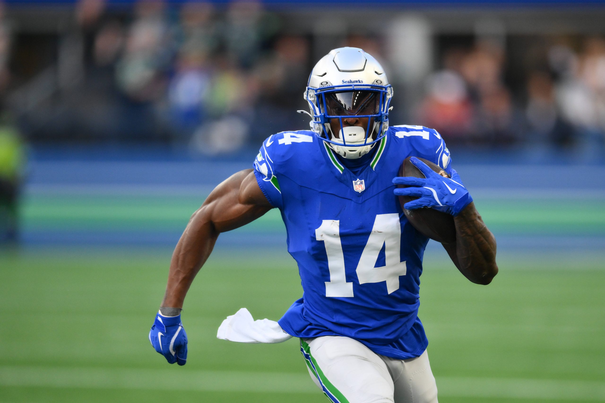 Ein Footballspieler, DK Metcalf, in einem blauen Trikot mit der Nummer 14 und einem silbernen Helm, sprintet über das Feld und hält den Ball mit seinem rechten Arm. Der unscharfe Hintergrund zeigt ein Publikum und Stadionlichter, was darauf schließen lässt, dass ein spannendes Seahawks-Spiel im Gange ist. Diese Beschreibung wurde mit der FootballR KI automatisch generiert.