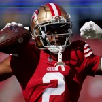 Deommodore Lenoir, ein Footballspieler in rotem Trikot und goldenem Helm, posiert triumphierend und hält einen Football in seiner rechten Hand. Sein linker Arm ist in stolzer Haltung vor dem Hintergrund eines verschwommenen Stadions voller Rottöne gestreckt und fängt die Essenz eines sonnigen Tages ein. Diese Beschreibung wurde mit der FootballR KI automatisch generiert.