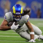 Spieltag 4 - Ein Footballspieler, Puka Nacua, in einem blau-gelben Helm und einer weißen Uniform, stürzt sich auf ein grünes Feld nach vorne und hält den Football fest in beiden Händen. Die Intensität ist sichtbar, als er sich streckt, um vor dem verschwommenen Hintergrund, der die Farben seines Teams widerspiegelt, zusätzliche Meter zu gewinnen. Diese Beschreibung wurde mit der FootballR KI automatisch generiert.