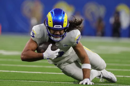 Spieltag 4 - Ein Footballspieler, Puka Nacua, in einem blau-gelben Helm und einer weißen Uniform, stürzt sich auf ein grünes Feld nach vorne und hält den Football fest in beiden Händen. Die Intensität ist sichtbar, als er sich streckt, um vor dem verschwommenen Hintergrund, der die Farben seines Teams widerspiegelt, zusätzliche Meter zu gewinnen. Diese Beschreibung wurde mit der FootballR KI automatisch generiert.