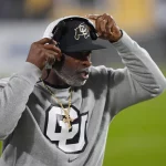 Deion Sanders auf einem Footballfeld trägt eine schwarze Kappe mit einem Logo, ein Headset und ein graues Sweatshirt mit einem großen „CU“-Logo. Er zeigt mit einer Hand auf sein Headset. Der Hintergrund ist unscharf und hebt grünes Gras und Stadionlichter hervor. Diese Beschreibung wurde mit der FootballR KI automatisch generiert.