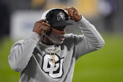 Deion Sanders auf einem Footballfeld trägt eine schwarze Kappe mit einem Logo, ein Headset und ein graues Sweatshirt mit einem großen „CU“-Logo. Er zeigt mit einer Hand auf sein Headset. Der Hintergrund ist unscharf und hebt grünes Gras und Stadionlichter hervor. Diese Beschreibung wurde mit der FootballR KI automatisch generiert.