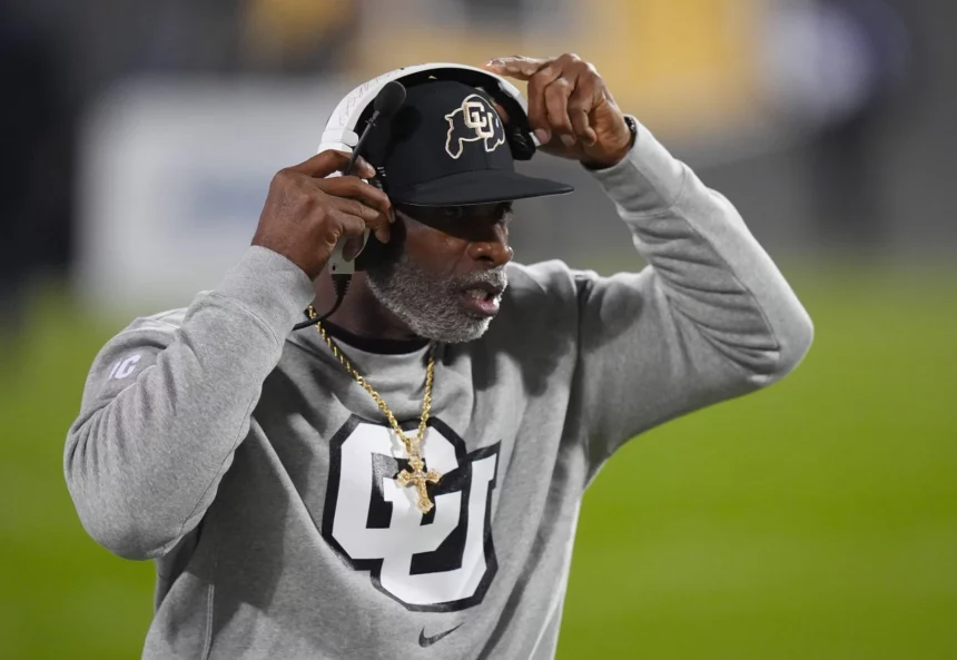 Deion Sanders auf einem Footballfeld trägt eine schwarze Kappe mit einem Logo, ein Headset und ein graues Sweatshirt mit einem großen „CU“-Logo. Er zeigt mit einer Hand auf sein Headset. Der Hintergrund ist unscharf und hebt grünes Gras und Stadionlichter hervor. Diese Beschreibung wurde mit der FootballR KI automatisch generiert.