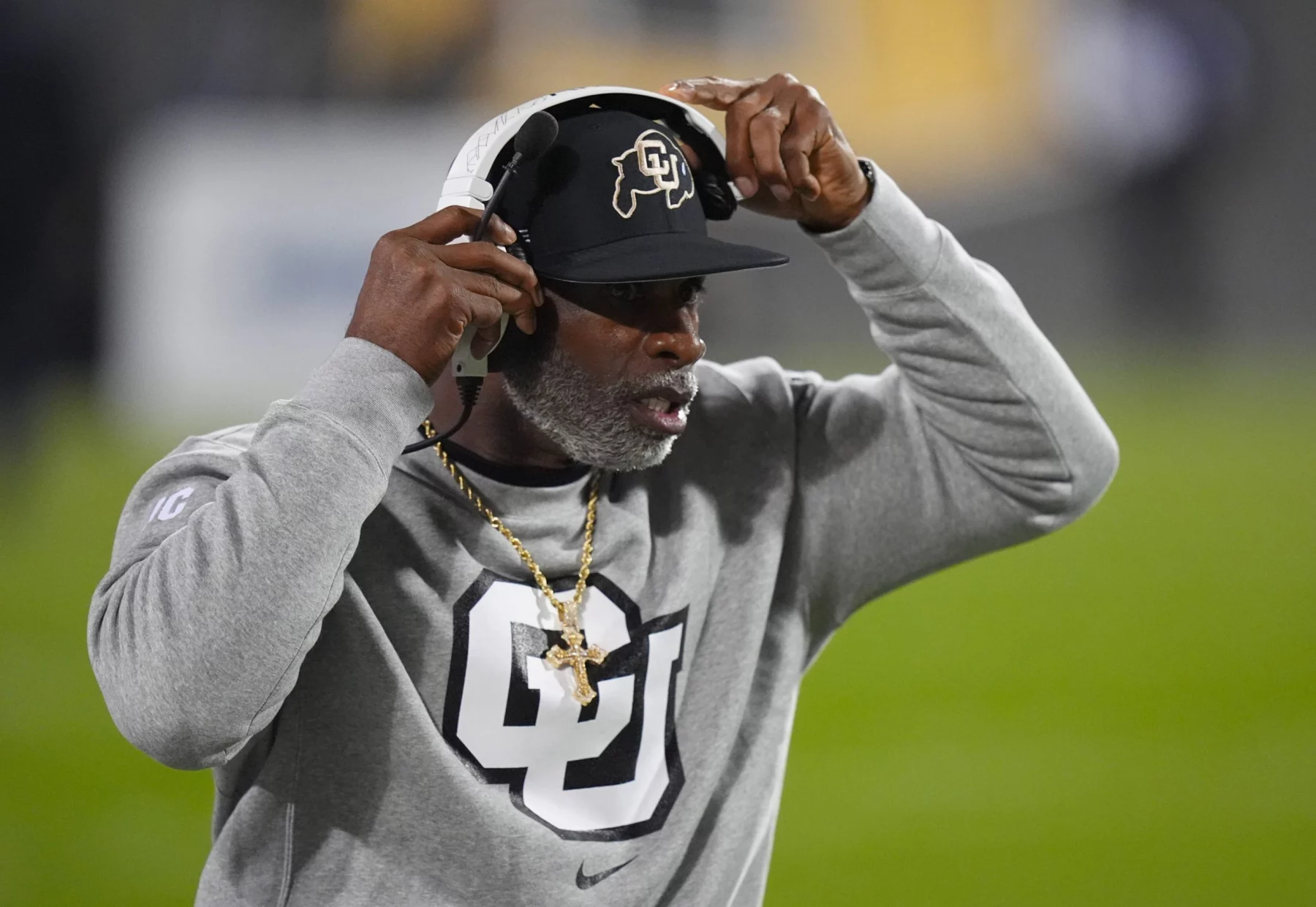 Deion Sanders auf einem Footballfeld trägt eine schwarze Kappe mit einem Logo, ein Headset und ein graues Sweatshirt mit einem großen „CU“-Logo. Er zeigt mit einer Hand auf sein Headset. Der Hintergrund ist unscharf und hebt grünes Gras und Stadionlichter hervor. Diese Beschreibung wurde mit der FootballR KI automatisch generiert.