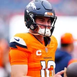 Fantasy Football Update: Bo Nix in orangefarbener Denver Broncos-Uniform und Helm lächelt auf dem Feld und verkörpert den Nervenkitzel des Fantasy Football. Er trägt einen Kapitänsaufnäher mit dem „C“-Symbol und das Trikot mit der Nummer 10 und steht zwischen verschwommenen Figuren und Zuschauern in einer Stadionkulisse, bereit für jede Playoff-Strategie. Diese Beschreibung wurde mit der FootballR KI automatisch generiert.