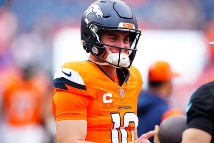 Fantasy Football Update: Bo Nix in orangefarbener Denver Broncos-Uniform und Helm lächelt auf dem Feld und verkörpert den Nervenkitzel des Fantasy Football. Er trägt einen Kapitänsaufnäher mit dem „C“-Symbol und das Trikot mit der Nummer 10 und steht zwischen verschwommenen Figuren und Zuschauern in einer Stadionkulisse, bereit für jede Playoff-Strategie. Diese Beschreibung wurde mit der FootballR KI automatisch generiert.