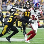 Darius Slayton, der Star-WR der Giants, weicht gekonnt einem Tackle eines gegnerischen Spielers in Schwarz und Gelb aus. In seiner weiß-roten Uniform hält er den Ball fest, während er den Verteidiger abwehrt. Die Action spielt sich auf einem Rasenplatz ab, im Hintergrund eine begeisterte Menschenmenge. Diese Beschreibung wurde mit der FootballR KI automatisch generiert.