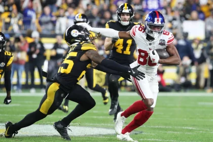 Darius Slayton, der Star-WR der Giants, weicht gekonnt einem Tackle eines gegnerischen Spielers in Schwarz und Gelb aus. In seiner weiß-roten Uniform hält er den Ball fest, während er den Verteidiger abwehrt. Die Action spielt sich auf einem Rasenplatz ab, im Hintergrund eine begeisterte Menschenmenge. Diese Beschreibung wurde mit der FootballR KI automatisch generiert.