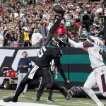New York Jets Receiver Garrett Wilson in schwarzer Uniform springt hoch, um mit einer Hand einen Pass zu fangen, während ein Spieler der Texans in weiß-blauer Uniform versucht, ihn zu blocken. Die Szene spielt in einem überfüllten Stadion mit jubelnden Zuschauern. Beide Sportler stehen in der Nähe der Seitenlinie, nahe der Endzone. Diese Beschreibung wurde mit der FootballR KI automatisch generiert.