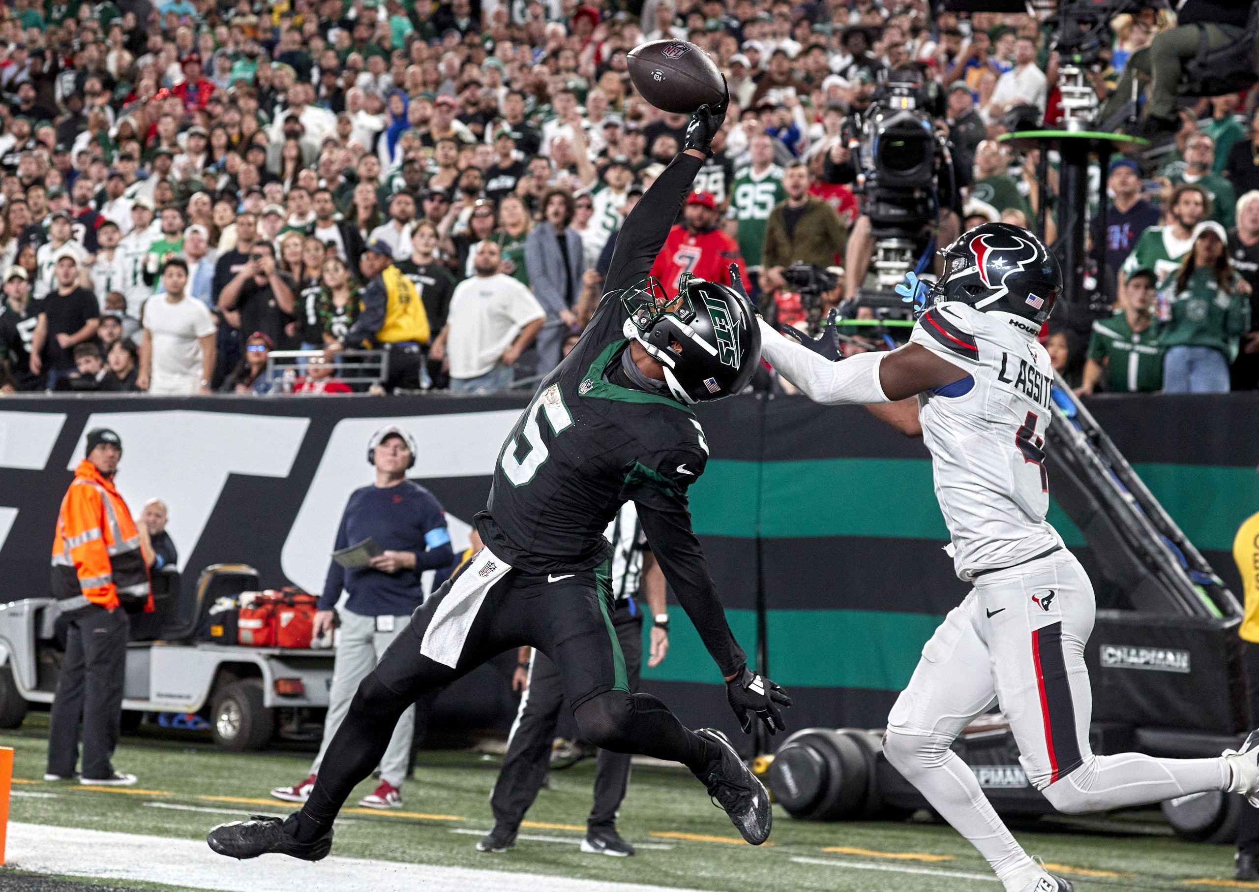 New York Jets Receiver Garrett Wilson in schwarzer Uniform springt hoch, um mit einer Hand einen Pass zu fangen, während ein Spieler der Texans in weiß-blauer Uniform versucht, ihn zu blocken. Die Szene spielt in einem überfüllten Stadion mit jubelnden Zuschauern. Beide Sportler stehen in der Nähe der Seitenlinie, nahe der Endzone. Diese Beschreibung wurde mit der FootballR KI automatisch generiert.