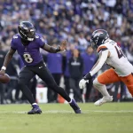 Lamar Jackson, im lila Trikot mit der Nummer 8, schießt gekonnt mit dem Ball, während ein anderer Spieler in weiß-orangefarbener Uniform zum Tackling eilt. Das grüne Spielfeld bildet einen Kontrast zur verschwommenen Zuschauermenge im Hintergrund. Diese Beschreibung wurde mit der FootballR KI automatisch generiert.