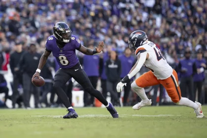 Lamar Jackson, im lila Trikot mit der Nummer 8, schießt gekonnt mit dem Ball, während ein anderer Spieler in weiß-orangefarbener Uniform zum Tackling eilt. Das grüne Spielfeld bildet einen Kontrast zur verschwommenen Zuschauermenge im Hintergrund. Diese Beschreibung wurde mit der FootballR KI automatisch generiert.