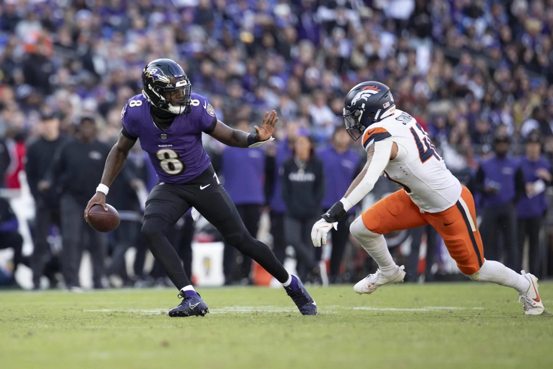 Lamar Jackson, im lila Trikot mit der Nummer 8, schießt gekonnt mit dem Ball, während ein anderer Spieler in weiß-orangefarbener Uniform zum Tackling eilt. Das grüne Spielfeld bildet einen Kontrast zur verschwommenen Zuschauermenge im Hintergrund. Diese Beschreibung wurde mit der FootballR KI automatisch generiert.