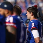 Daniel Jones in einem blauen Trikot mit der Nummer 8 steht mit der Hand auf dem Herzen an der Seitenlinie. Inmitten ähnlich gekleideter Giants-Teamkollegen erinnert die Szene an eine Zeremonie vor dem Spiel. Die verschwommene Stadionkulisse steigert die Vorfreude auf die Leistung von Daniel Jones. Diese Beschreibung wurde mit der FootballR KI automatisch generiert.