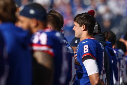 Daniel Jones in einem blauen Trikot mit der Nummer 8 steht mit der Hand auf dem Herzen an der Seitenlinie. Inmitten ähnlich gekleideter Giants-Teamkollegen erinnert die Szene an eine Zeremonie vor dem Spiel. Die verschwommene Stadionkulisse steigert die Vorfreude auf die Leistung von Daniel Jones. Diese Beschreibung wurde mit der FootballR KI automatisch generiert.