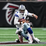 Dak Prescott im weißen Trikot mit der Nummer 4, der mit dem Ball rennt, wird auf einem Footballfeld von einem Gegner im schwarzen Trikot angegriffen. In der Nähe der 50-Yard-Linie, wo das große Falcons-Logo ein Spiel der Atlanta Falcons markiert, steht Dak Prescott nach einer unerwarteten Oberschenkelverletzung vor seiner Herausforderung der Saison. Diese Beschreibung wurde mit der FootballR KI automatisch generiert.
