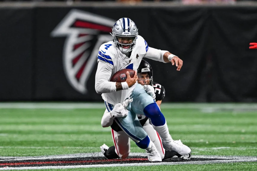 Dak Prescott im weißen Trikot mit der Nummer 4, der mit dem Ball rennt, wird auf einem Footballfeld von einem Gegner im schwarzen Trikot angegriffen. In der Nähe der 50-Yard-Linie, wo das große Falcons-Logo ein Spiel der Atlanta Falcons markiert, steht Dak Prescott nach einer unerwarteten Oberschenkelverletzung vor seiner Herausforderung der Saison. Diese Beschreibung wurde mit der FootballR KI automatisch generiert.