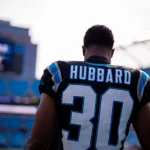 Chuba Hubbard in einem Carolina Panthers-Trikot mit der Nummer „30“ steht auf dem Spielfeld. Hinter ihm erstreckt sich das Stadion und zeigt Reihen leerer blauer Sitze sowie eine elektronische Anzeigetafel, auf der Spieldetails angezeigt werden. Der Spieler ist mit dem Rücken zur Kamera gerichtet. Diese Beschreibung wurde mit der FootballR KI automatisch generiert.