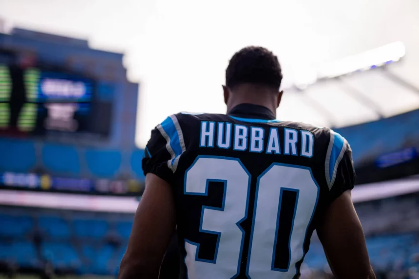 Chuba Hubbard in einem Carolina Panthers-Trikot mit der Nummer „30“ steht auf dem Spielfeld. Hinter ihm erstreckt sich das Stadion und zeigt Reihen leerer blauer Sitze sowie eine elektronische Anzeigetafel, auf der Spieldetails angezeigt werden. Der Spieler ist mit dem Rücken zur Kamera gerichtet. Diese Beschreibung wurde mit der FootballR KI automatisch generiert.