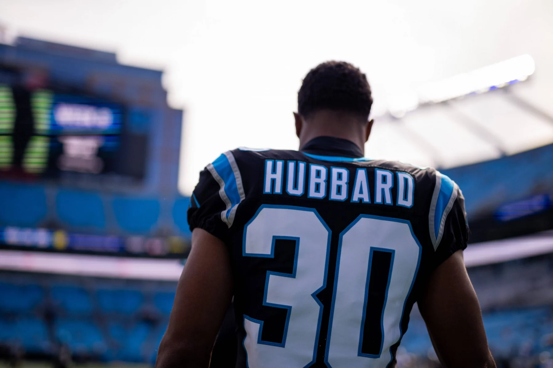Chuba Hubbard in einem Carolina Panthers-Trikot mit der Nummer „30“ steht auf dem Spielfeld. Hinter ihm erstreckt sich das Stadion und zeigt Reihen leerer blauer Sitze sowie eine elektronische Anzeigetafel, auf der Spieldetails angezeigt werden. Der Spieler ist mit dem Rücken zur Kamera gerichtet. Diese Beschreibung wurde mit der FootballR KI automatisch generiert.