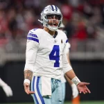Dak Prescott in einem weißen Trikot mit blauen Akzenten und der Nummer 4, steht auf dem Feld und blickt nach vorne, als würde er schreien. Seine Teamkollegen sind hinter ihm verschwommen. Das Sternlogo auf dem Helm zeigt, dass er zu den Dallas Cowboys gehört, obwohl er derzeit auf der Verletztenliste steht. Diese Beschreibung wurde mit der FootballR KI automatisch generiert.