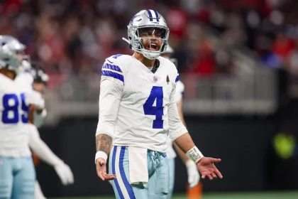 Dak Prescott in einem weißen Trikot mit blauen Akzenten und der Nummer 4, steht auf dem Feld und blickt nach vorne, als würde er schreien. Seine Teamkollegen sind hinter ihm verschwommen. Das Sternlogo auf dem Helm zeigt, dass er zu den Dallas Cowboys gehört, obwohl er derzeit auf der Verletztenliste steht. Diese Beschreibung wurde mit der FootballR KI automatisch generiert.