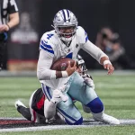 Dak Prescott in einer weiß-blauen Dallas Cowboys-Uniform, Nummer 4, läuft mit dem Ball, während er von einem Gegner in einer schwarz-roten Uniform angegriffen wird. Die Aktion findet auf einem grünen Feld mit einem Schiedsrichter im Hintergrund statt. Der Spieler scheint darauf konzentriert zu sein, das Spiel voranzutreiben. Diese Beschreibung wurde mit der FootballR KI automatisch generiert.