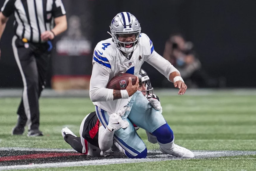 Dak Prescott in einer weiß-blauen Dallas Cowboys-Uniform, Nummer 4, läuft mit dem Ball, während er von einem Gegner in einer schwarz-roten Uniform angegriffen wird. Die Aktion findet auf einem grünen Feld mit einem Schiedsrichter im Hintergrund statt. Der Spieler scheint darauf konzentriert zu sein, das Spiel voranzutreiben. Diese Beschreibung wurde mit der FootballR KI automatisch generiert.