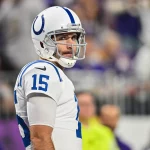 Joe Flacco in weiß-blauer Uniform steht mit der Nummer 15 auf dem Feld. Mit seinem klaren Visier und dem konzentrierten Gesichtsausdruck ist er eine Schlüsselfigur der Colts. Im Hintergrund sitzen verschwommene Zuschauer unter hellem Stadionlicht. Diese Beschreibung wurde mit der FootballR KI automatisch generiert.