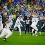 NFL Week 9: In einem spannenden Footballspiel der 9. Woche wirft ein Spieler in blau-weißer Uniform einen Pass. Teamkollegen und Gegner, in Gelb und Grün gekleidet, manövrieren auf dem Feld. Der Schiedsrichter beobachtet im Hintergrund, während die Zuschauer in den unterschiedlichsten Farben die Tribünen unter bewölktem Himmel füllen. Diese Beschreibung wurde mit der FootballR KI automatisch generiert.