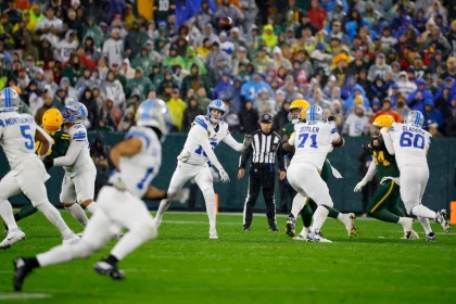 NFL Week 9: In einem spannenden Footballspiel der 9. Woche wirft ein Spieler in blau-weißer Uniform einen Pass. Teamkollegen und Gegner, in Gelb und Grün gekleidet, manövrieren auf dem Feld. Der Schiedsrichter beobachtet im Hintergrund, während die Zuschauer in den unterschiedlichsten Farben die Tribünen unter bewölktem Himmel füllen. Diese Beschreibung wurde mit der FootballR KI automatisch generiert.