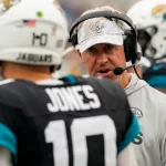Ein Footballtrainer, Doug Pederson, trägt ein Headset und eine Mannschaftskappe und spricht mit einem Spieler in einer Jaguars-Uniform mit „Jones“ und der Nummer 10 auf dem Rücken. Der Spieler trägt einen weißen Helm, während im verschwommenen Hintergrund auf dem Sportplatz weitere behelmte Teamkollegen zu sehen sind. Diese Beschreibung wurde mit der FootballR KI automatisch generiert.