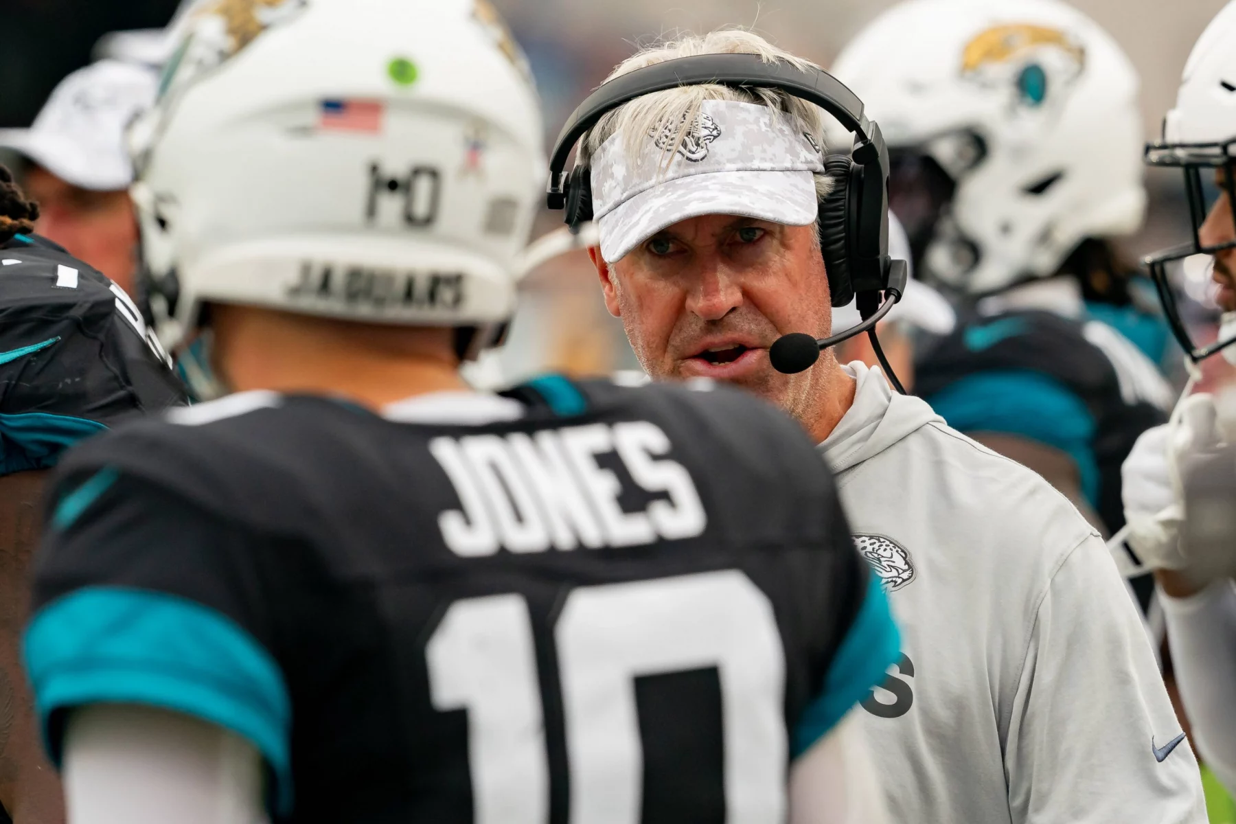 Ein Footballtrainer, Doug Pederson, trägt ein Headset und eine Mannschaftskappe und spricht mit einem Spieler in einer Jaguars-Uniform mit „Jones“ und der Nummer 10 auf dem Rücken. Der Spieler trägt einen weißen Helm, während im verschwommenen Hintergrund auf dem Sportplatz weitere behelmte Teamkollegen zu sehen sind. Diese Beschreibung wurde mit der FootballR KI automatisch generiert.