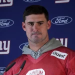 Ein Footballspieler in rotem Trikot und grauem Kapuzenpullover, Daniel Jones, steht vor einem Hintergrund, der mit Logos der New York Giants und Ford geschmückt ist. Er erscheint bei einer Pressekonferenz und behält einen ernsten Gesichtsausdruck bei, während Mikrofone ihn umgeben. Diese Beschreibung wurde mit der FootballR KI automatisch generiert.