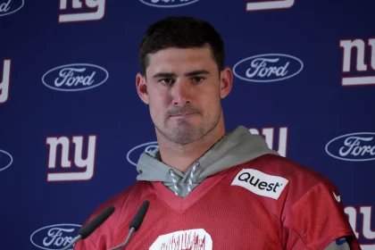 Ein Footballspieler in rotem Trikot und grauem Kapuzenpullover, Daniel Jones, steht vor einem Hintergrund, der mit Logos der New York Giants und Ford geschmückt ist. Er erscheint bei einer Pressekonferenz und behält einen ernsten Gesichtsausdruck bei, während Mikrofone ihn umgeben. Diese Beschreibung wurde mit der FootballR KI automatisch generiert.