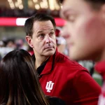 Ein Mann in einer roten Jacke mit einem weißen „IU“-Logo blickt in einem überfüllten Veranstaltungsort nach links. Inmitten der lebhaften Atmosphäre, in der Menschen in Rot und verschwommene Figuren, darunter ein Polizist, den Hintergrund unter hellen Lichtern füllen, kann man Curt Cignetti sehen, der zur Energie beiträgt. Diese Beschreibung wurde mit der FootballR KI automatisch generiert.