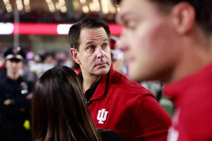 Ein Mann in einer roten Jacke mit einem weißen „IU“-Logo blickt in einem überfüllten Veranstaltungsort nach links. Inmitten der lebhaften Atmosphäre, in der Menschen in Rot und verschwommene Figuren, darunter ein Polizist, den Hintergrund unter hellen Lichtern füllen, kann man Curt Cignetti sehen, der zur Energie beiträgt. Diese Beschreibung wurde mit der FootballR KI automatisch generiert.