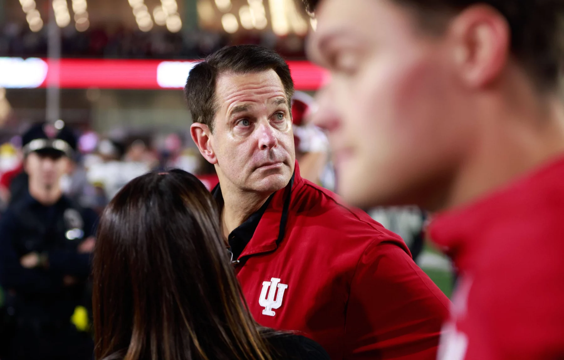 Ein Mann in einer roten Jacke mit einem weißen „IU“-Logo blickt in einem überfüllten Veranstaltungsort nach links. Inmitten der lebhaften Atmosphäre, in der Menschen in Rot und verschwommene Figuren, darunter ein Polizist, den Hintergrund unter hellen Lichtern füllen, kann man Curt Cignetti sehen, der zur Energie beiträgt. Diese Beschreibung wurde mit der FootballR KI automatisch generiert.