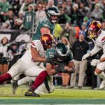 Ein Moment aus einem Footballspiel zeigt Saquon Barkley in einem grünen Trikot, der von zwei Spielern in weißen Trikots angegriffen wird. Die Fans des grünen Teams sind im Hintergrund zu sehen und beide Teams tragen Helme. Die Action ist intensiv, wobei ein Tackler nach dem Ball greift. Diese Beschreibung wurde mit der FootballR KI automatisch generiert.