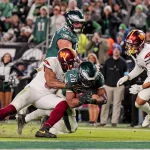 Ein Moment aus einem Footballspiel zeigt Saquon Barkley in einem grünen Trikot, der von zwei Spielern in weißen Trikots angegriffen wird. Die Fans des grünen Teams sind im Hintergrund zu sehen und beide Teams tragen Helme. Die Action ist intensiv, wobei ein Tackler nach dem Ball greift. Diese Beschreibung wurde mit der FootballR KI automatisch generiert.