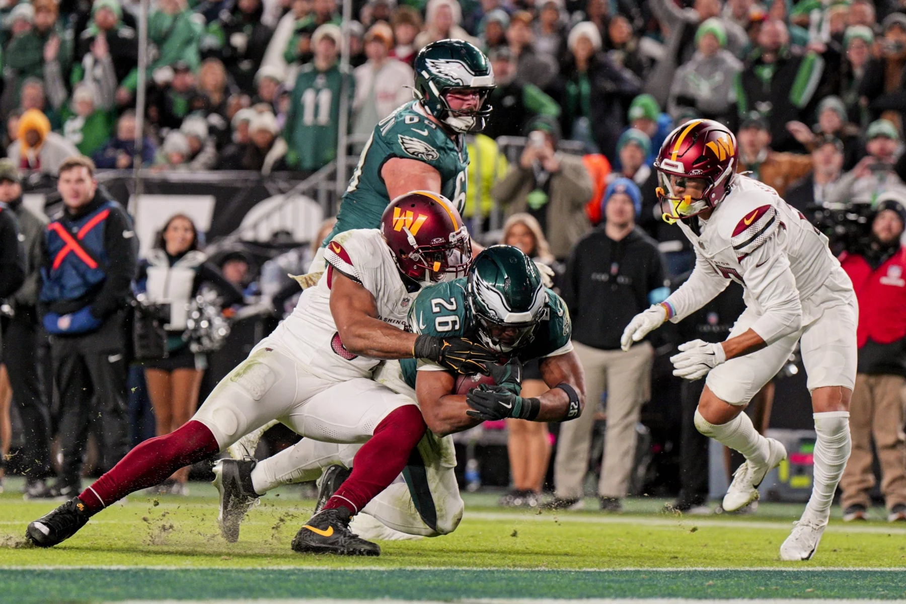 Ein Moment aus einem Footballspiel zeigt Saquon Barkley in einem grünen Trikot, der von zwei Spielern in weißen Trikots angegriffen wird. Die Fans des grünen Teams sind im Hintergrund zu sehen und beide Teams tragen Helme. Die Action ist intensiv, wobei ein Tackler nach dem Ball greift. Diese Beschreibung wurde mit der FootballR KI automatisch generiert.