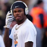 Travis Hunter, ein Footballspieler in weißem Hemd und schwarzem Stirnband, rückt seine Kopfhörer zurecht. Um seinen Hals glänzt eine goldene Kette, er trägt weiße Handschuhe. Der unscharfe Hintergrund zeigt andere in der Nähe, darunter jemanden in einer orangefarbenen Warnweste. Diese Beschreibung wurde mit der FootballR KI automatisch generiert.