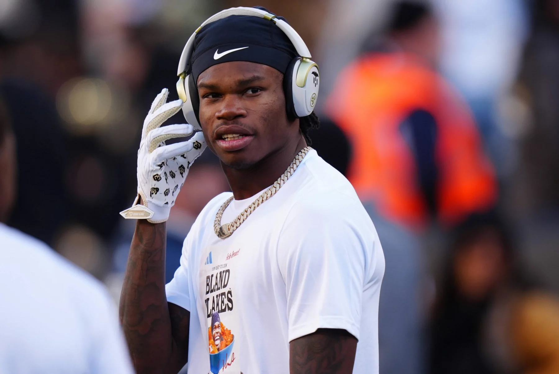 Travis Hunter, ein Footballspieler in weißem Hemd und schwarzem Stirnband, rückt seine Kopfhörer zurecht. Um seinen Hals glänzt eine goldene Kette, er trägt weiße Handschuhe. Der unscharfe Hintergrund zeigt andere in der Nähe, darunter jemanden in einer orangefarbenen Warnweste. Diese Beschreibung wurde mit der FootballR KI automatisch generiert.