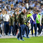 Mack Brown, der einen Kapuzenpullover mit Camouflage-Muster und der Aufschrift „UNC“ trägt, schreit leidenschaftlich am Spielfeldrand. Er ist von aufgeregten Teammitarbeitern und Spielern in Blau und Weiß umgeben. Im Hintergrund ist ein überfülltes Stadion mit verschwommenen Fans zu sehen. Im Vordergrund ist das Grasfeld zu sehen. Diese Beschreibung wurde mit der FootballR KI automatisch generiert.