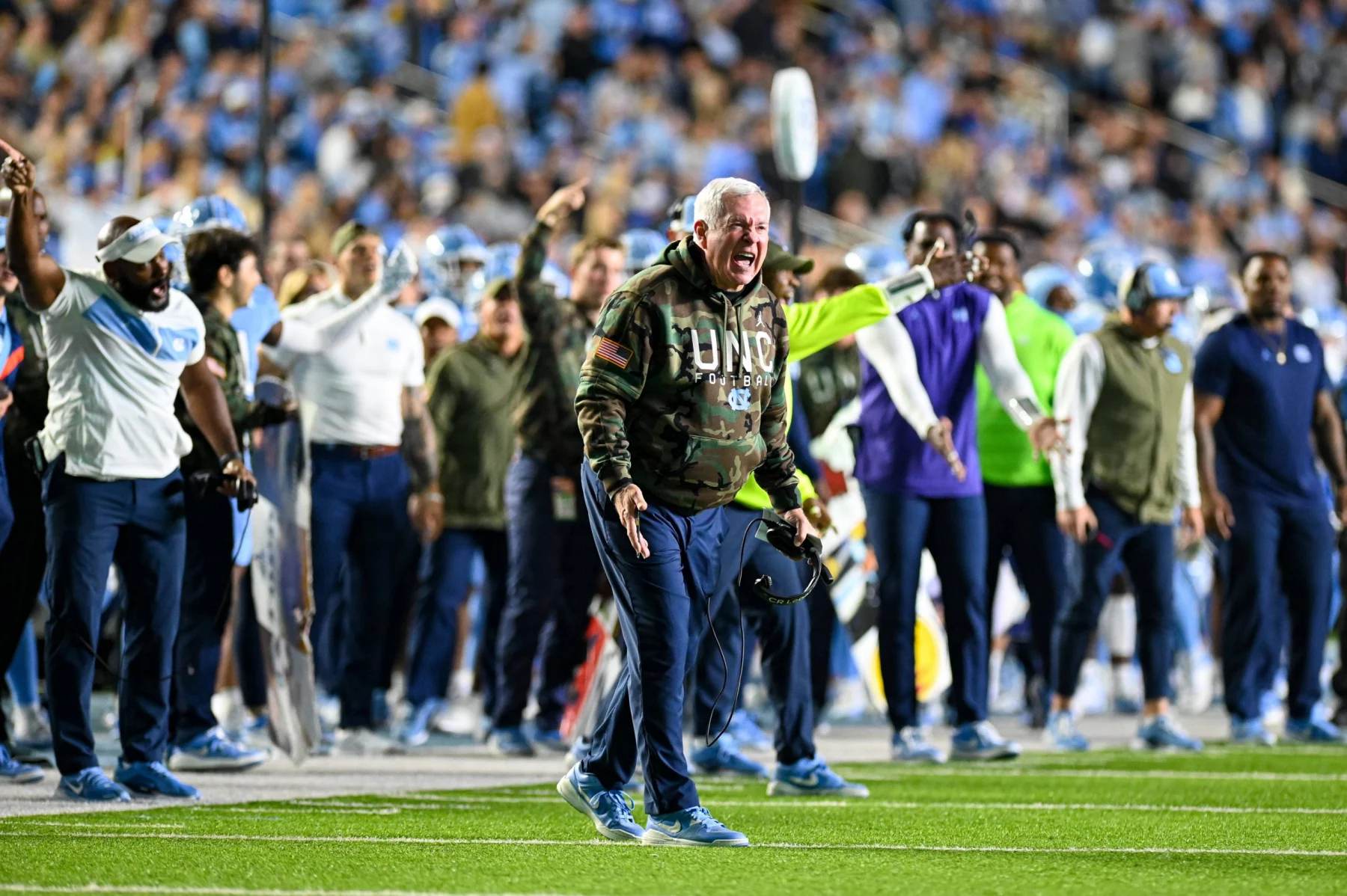 Mack Brown, der einen Kapuzenpullover mit Camouflage-Muster und der Aufschrift „UNC“ trägt, schreit leidenschaftlich am Spielfeldrand. Er ist von aufgeregten Teammitarbeitern und Spielern in Blau und Weiß umgeben. Im Hintergrund ist ein überfülltes Stadion mit verschwommenen Fans zu sehen. Im Vordergrund ist das Grasfeld zu sehen. Diese Beschreibung wurde mit der FootballR KI automatisch generiert.