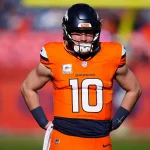 Bo Nix in orange-blauer Uniform mit der Nummer 10 steht in Woche 11 mit den Händen in den Hüften auf dem Feld. Er trägt einen Helm und hat einen konzentrierten Gesichtsausdruck. Im Hintergrund ist ein verschwommenes Stadion mit einer undeutlichen Menschenmenge und roten Bannern zu sehen. Diese Beschreibung wurde mit der FootballR KI automatisch generiert.