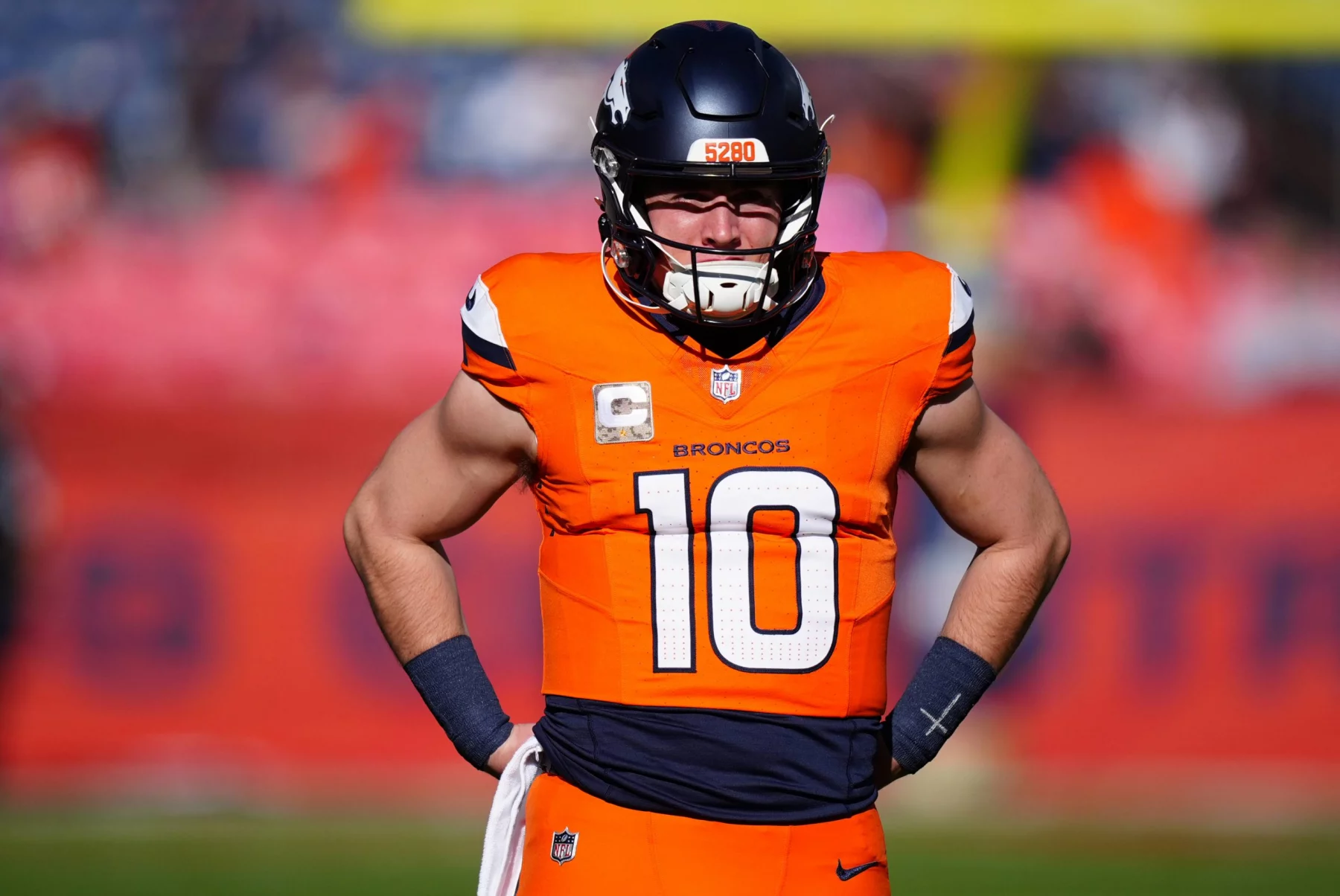 Bo Nix in orange-blauer Uniform mit der Nummer 10 steht in Woche 11 mit den Händen in den Hüften auf dem Feld. Er trägt einen Helm und hat einen konzentrierten Gesichtsausdruck. Im Hintergrund ist ein verschwommenes Stadion mit einer undeutlichen Menschenmenge und roten Bannern zu sehen. Diese Beschreibung wurde mit der FootballR KI automatisch generiert.