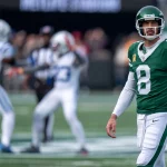 NFL Legenden: Aaron Rodgers in grüner Uniform mit der Nummer 8 steht auf dem Feld und blickt nach vorne. Im Hintergrund sind zwei gegnerische Spieler in weiß-blauen Uniformen zu sehen, die in Bewegung zu sein scheinen. Die Kulisse ist ein Freiluftstadion mit verschwommenem Publikum. Diese Beschreibung wurde mit der FootballR KI automatisch generiert.