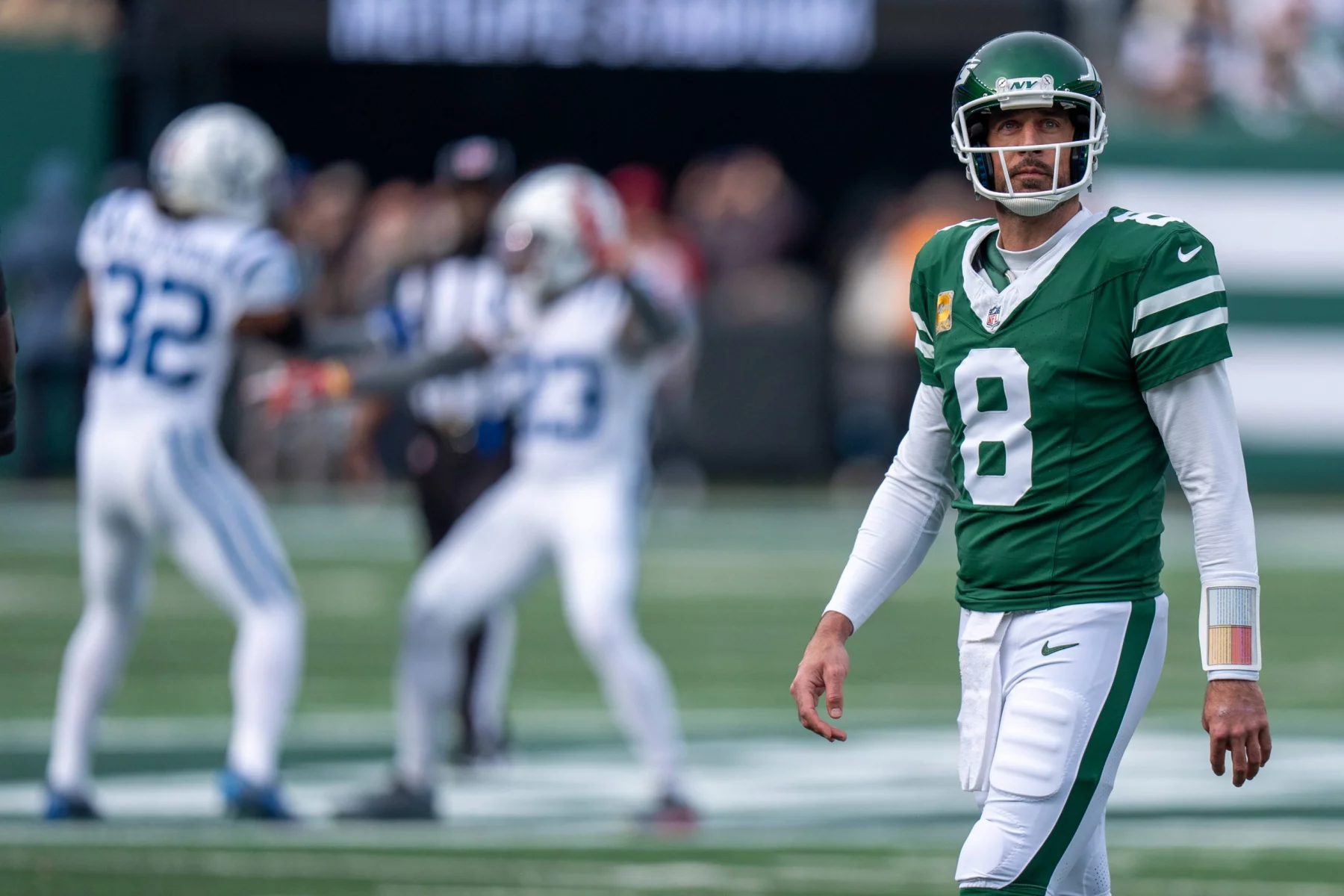 NFL Legenden: Aaron Rodgers in grüner Uniform mit der Nummer 8 steht auf dem Feld und blickt nach vorne. Im Hintergrund sind zwei gegnerische Spieler in weiß-blauen Uniformen zu sehen, die in Bewegung zu sein scheinen. Die Kulisse ist ein Freiluftstadion mit verschwommenem Publikum. Diese Beschreibung wurde mit der FootballR KI automatisch generiert.