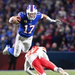NFL Playoffs: Josh Allen im blauen Trikot mit der Nummer 17 springt über einen Gegenspieler der gegnerischen Mannschaft in Rot und Weiß. Der Spieler hält den Ball fest im Griff, aufgenommen mitten im Flug vor einem verschwommenen Hintergrund begeisterter Fans. Diese Beschreibung wurde mit der FootballR KI automatisch generiert.